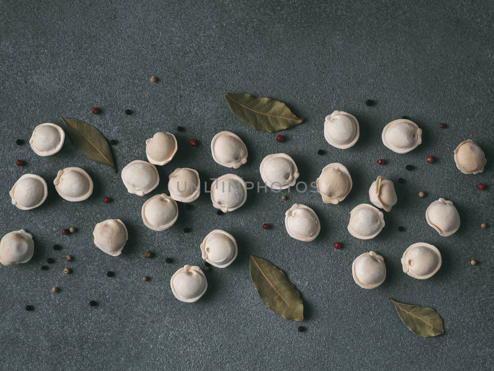 Pattern of frozen uncooked russian pelmeni with peppercorns and bay leaves on black background. Creative layout of dumplings. Beautiful scattered raw dumplings. Top view or flat lay.