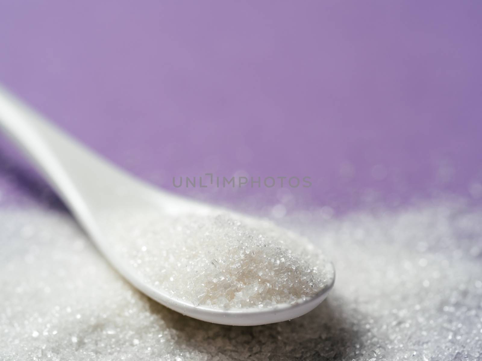 Sugar in white ceramic spoon on lilac background. Spoon with white sugar on violet background, closeup. Shallow DOF. Copy space for text.