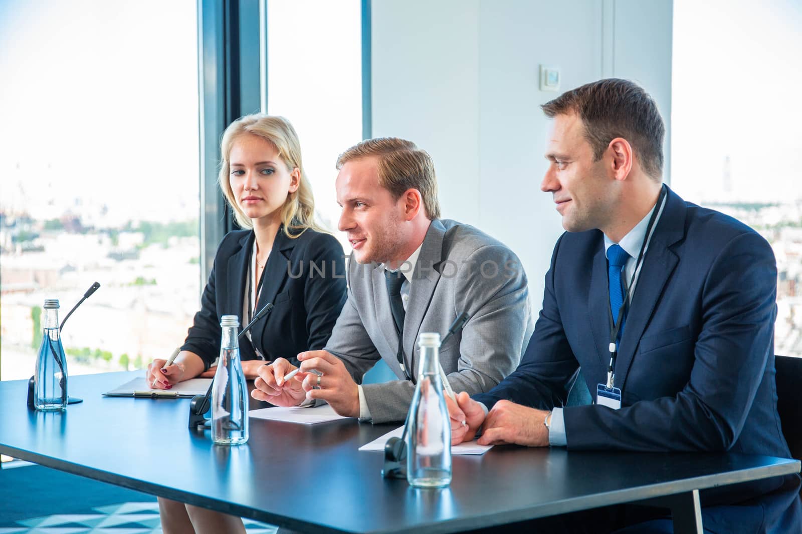 Row of speakers sitting in row by table in front of microphones while taking part in business conference