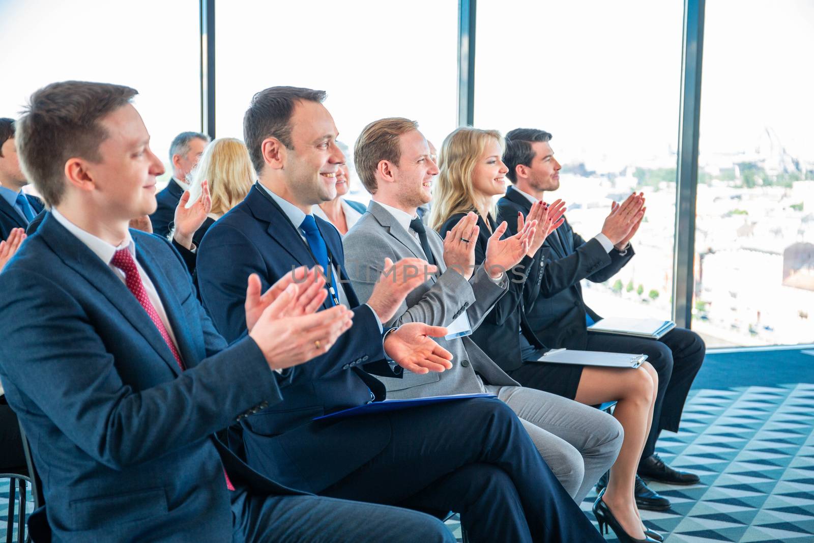 Group of happy business people in formalwear sitting at the chairs in conference hall and applauding to speaker