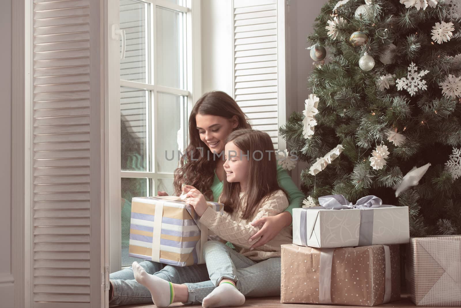 Happy mother and her little daughter unpack gift box near the Christmas tree at home