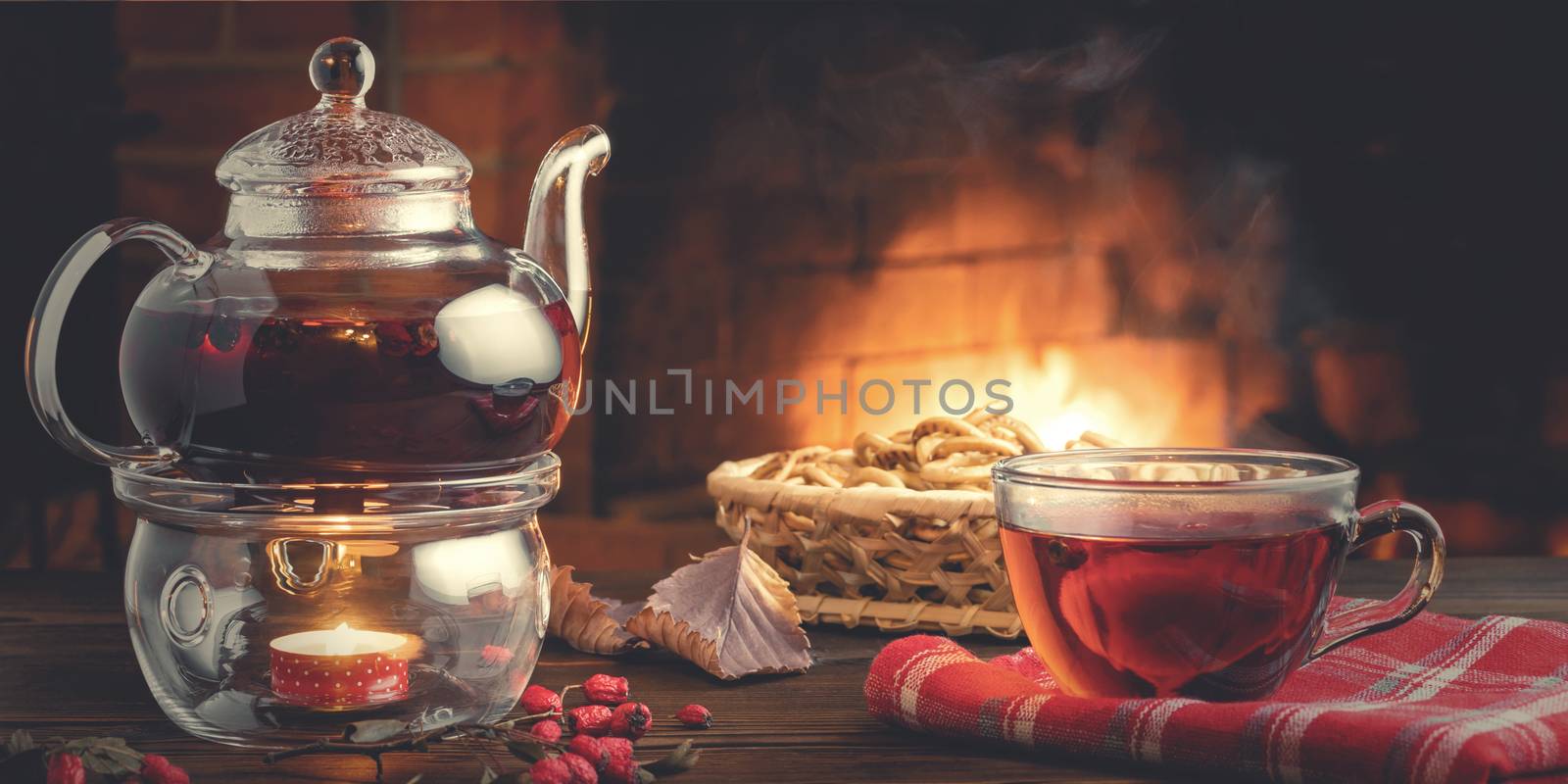 Tea with hawthorn in a glass cup and teapot with a tealight on a wooden table in a room with a burning fireplace by galsand