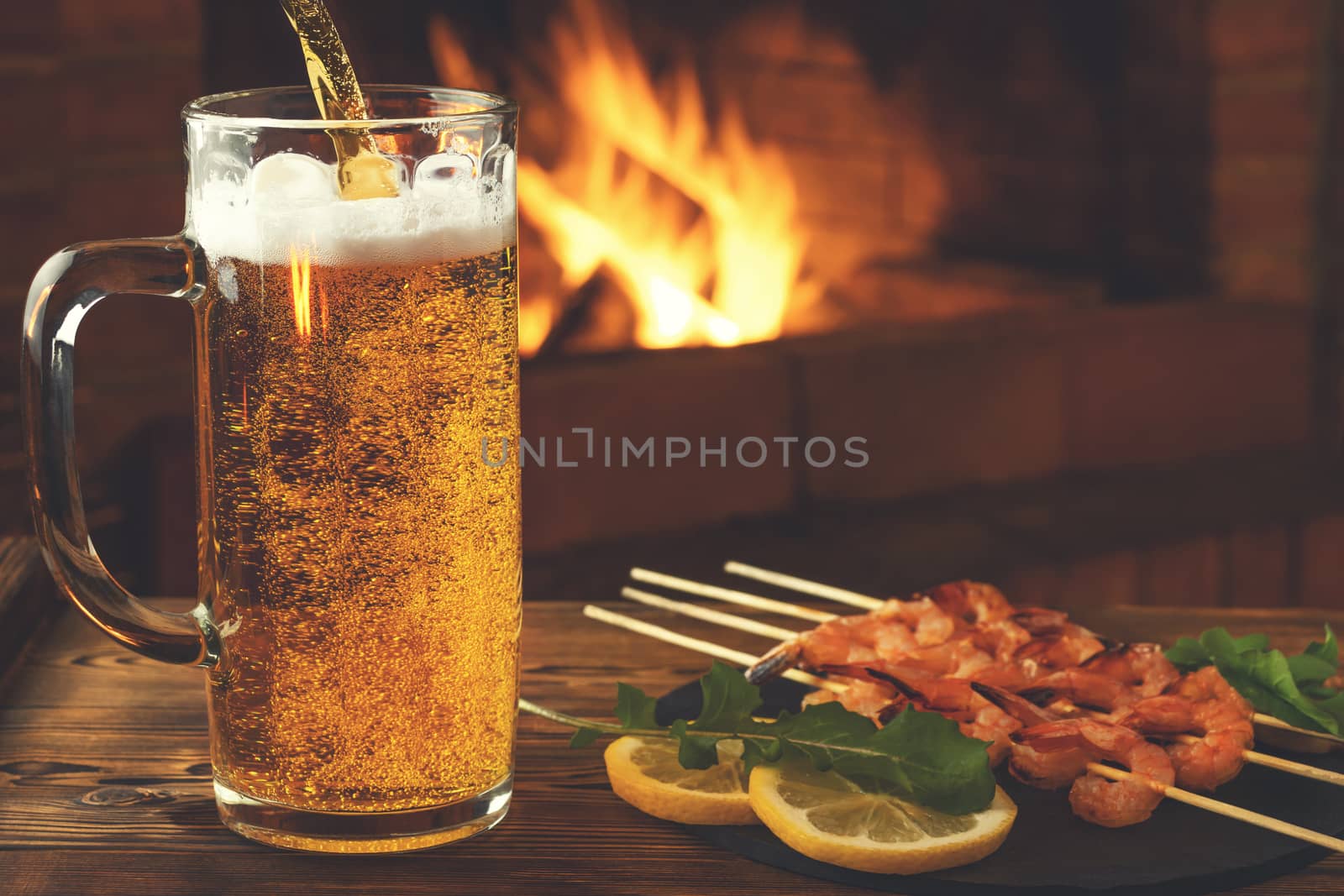 Pouring beer into a mug, shrimp fried on skewers with arugula, lemon on a black slate dish on a wooden table in a room with a fireplace by galsand