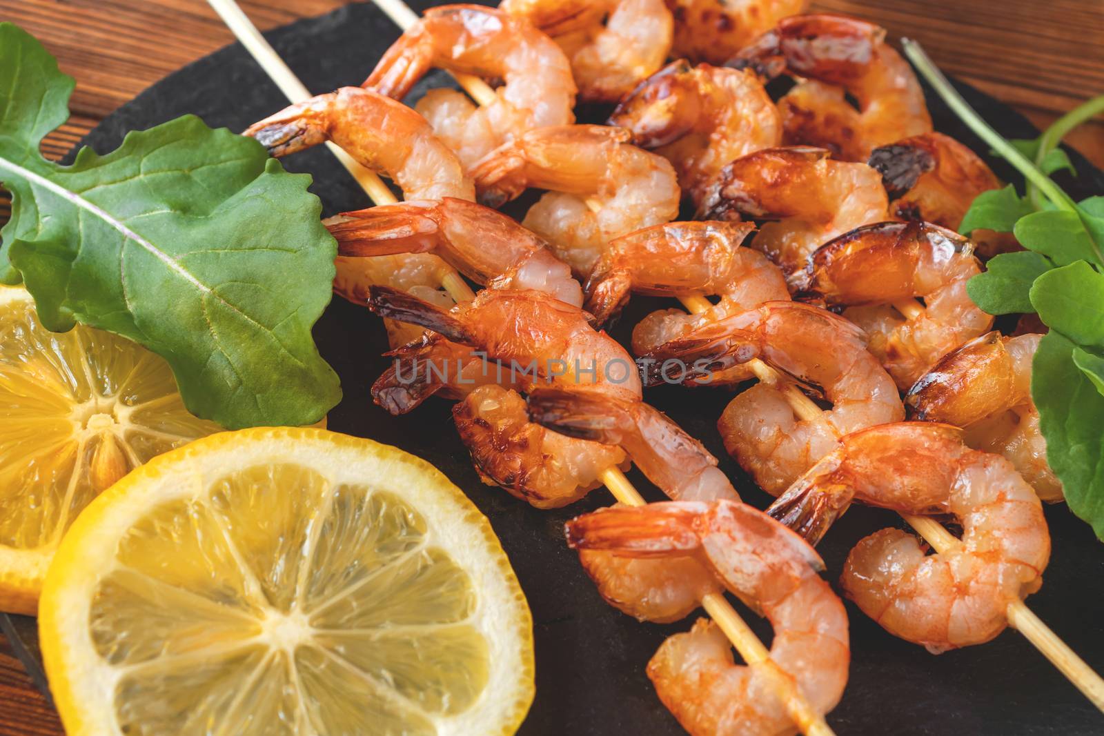 Shrimp fried on skewers with arugula and lemon on a black slate dish on a wooden table, top view, flat lay, close up by galsand