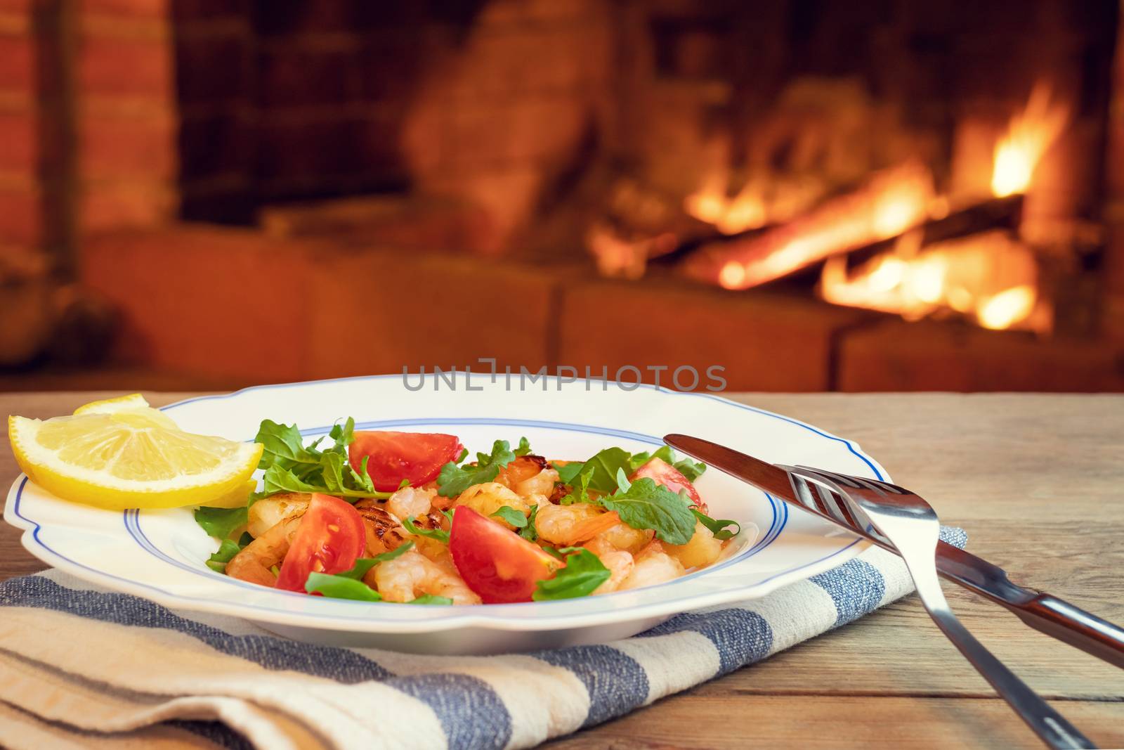 Fresh homemade salad of shrimp, arugula and tomato in a white plate on a wooden table in a room with a fireplace, close-up.