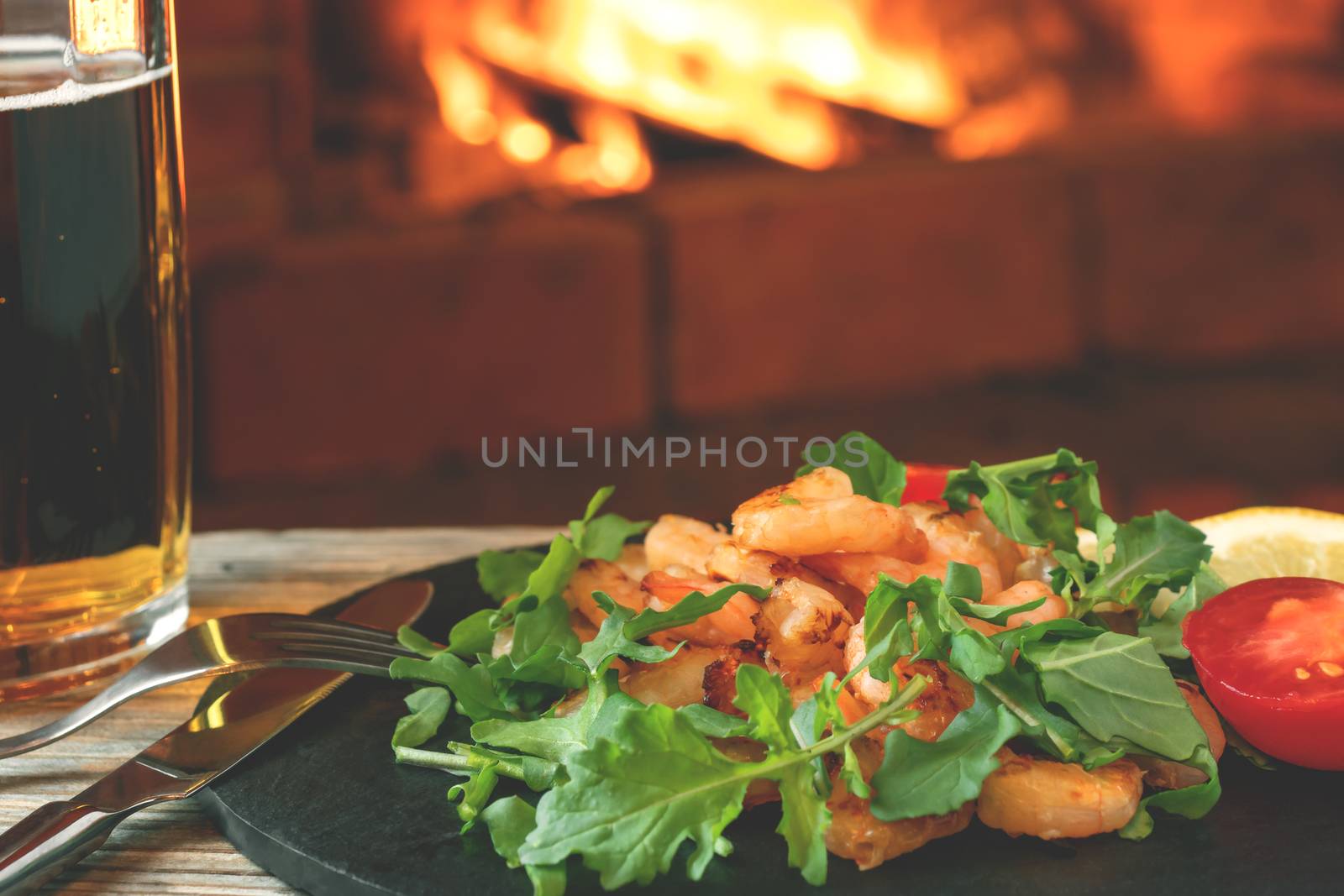 Fresh homemade salad of shrimp, arugula and tomato on a black slate dish and beer on a wooden table in a room with a fireplace, close-up by galsand