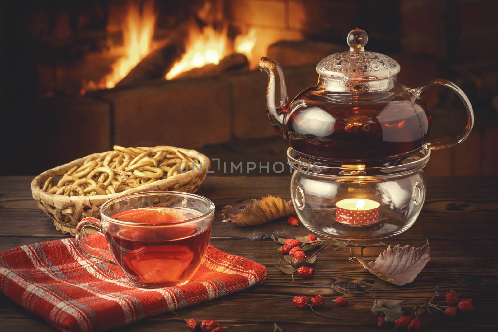 Tea with hawthorn in a glass cup and teapot with a tealight on a wooden table in a room with a burning fireplace.