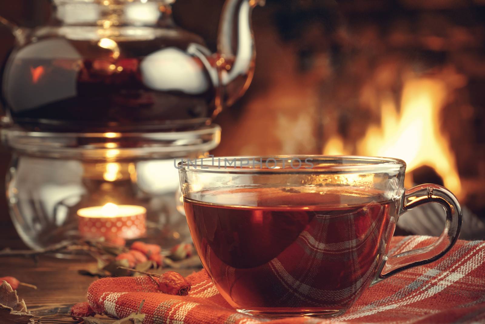 Tea with hawthorn in a glass cup and teapot with a tealight on a wooden table in a room with a burning fireplace by galsand