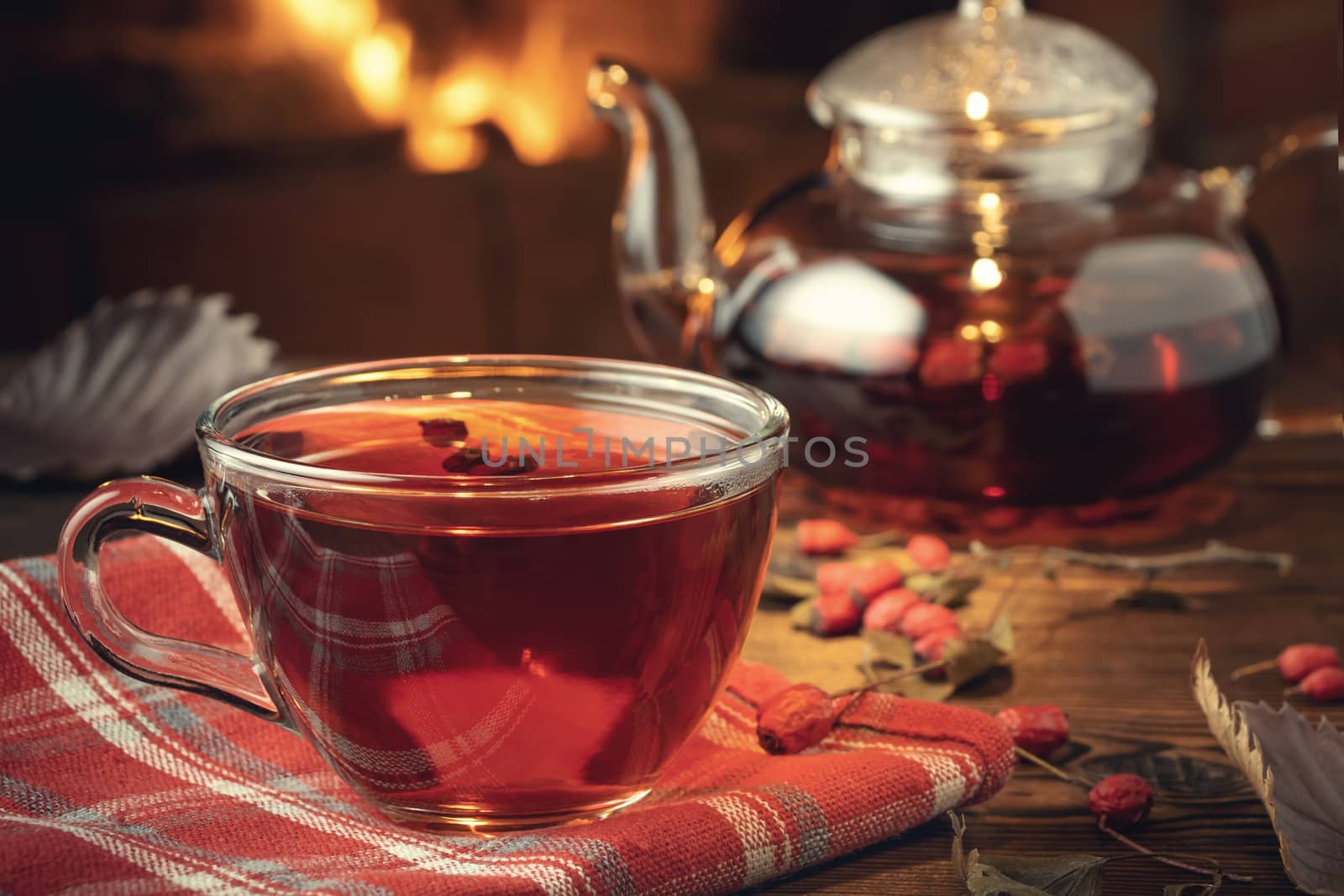 Tea with hawthorn in a glass cup and teapot on a wooden table in a room with a burning fireplace, closeup by galsand