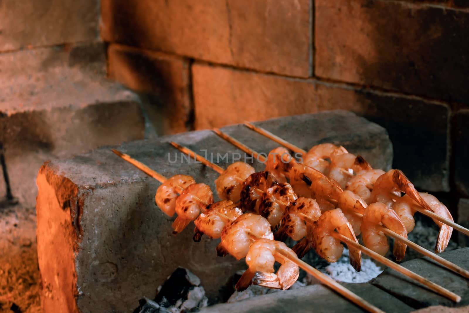 Shrimp frying on bamboo skewers on hot coals in a fireplace.