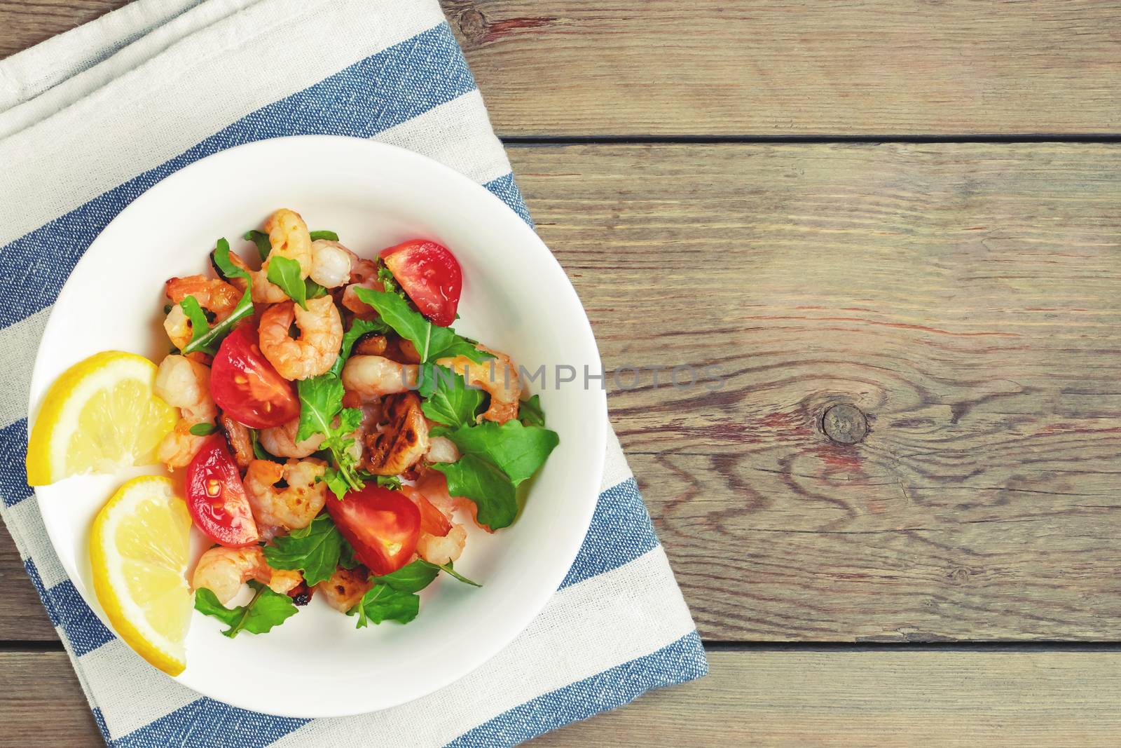 Fresh homemade salad of shrimp, arugula and tomato in a white plate on a wooden table, top view, flat lay, copyspace.