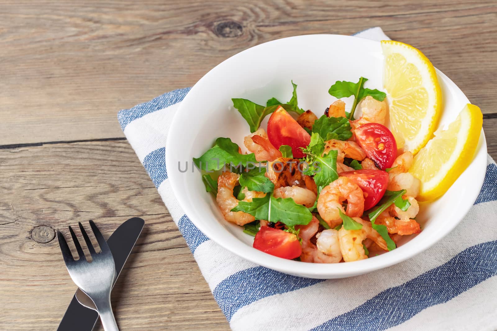 Fresh homemade salad of shrimp, arugula and tomato in a white plate on a wooden table.