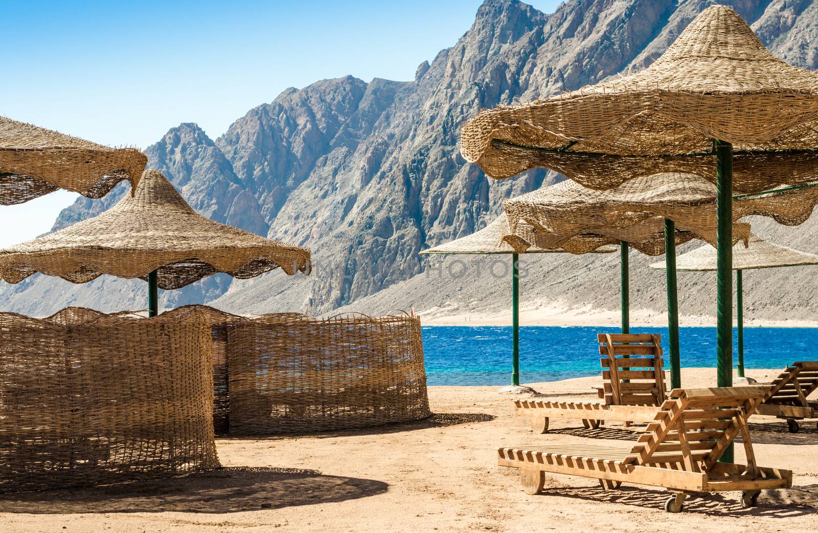 beach umbrellas and wooden lounge chairs on the sand of the beach against the backdrop of the sea and high rocky mountains