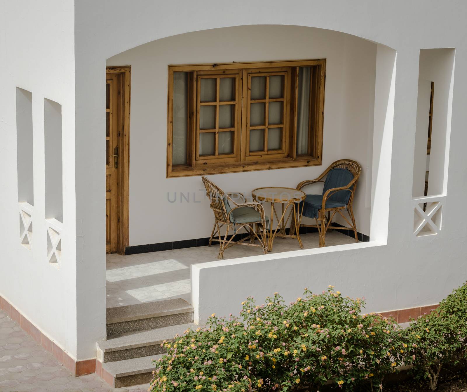 terrace of the resort house with chairs and a round table and a green bush nearby