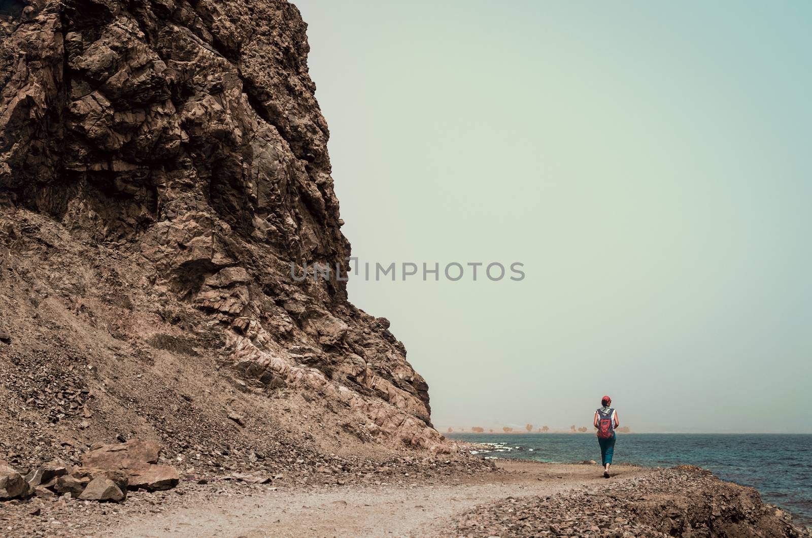 young girl tourist in a red cap with a backpack goes along the rocky shore of the Red Sea in Egypt