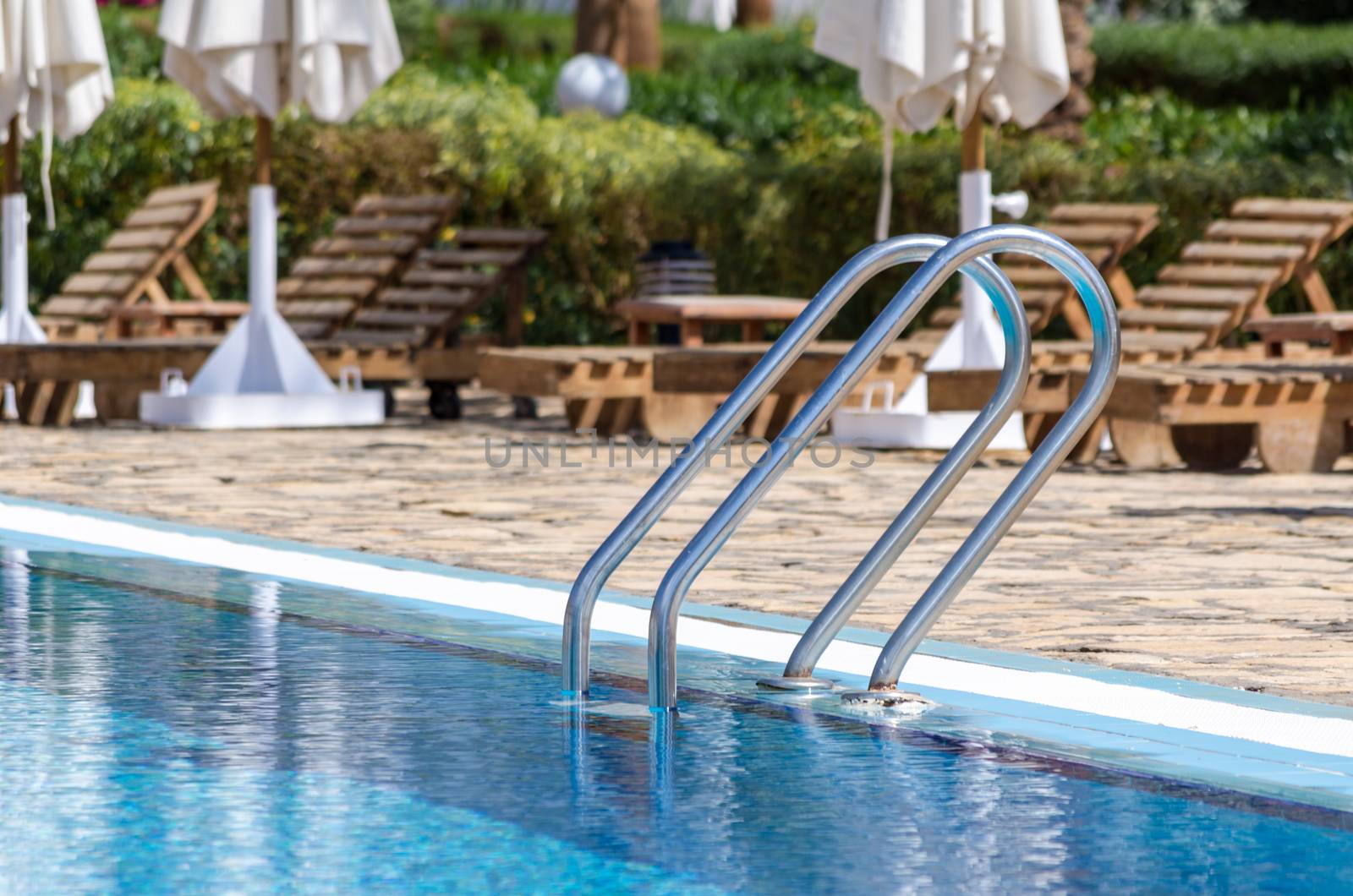 handrails by the pool with blue water on the background of sun loungers and folded white umbrellas