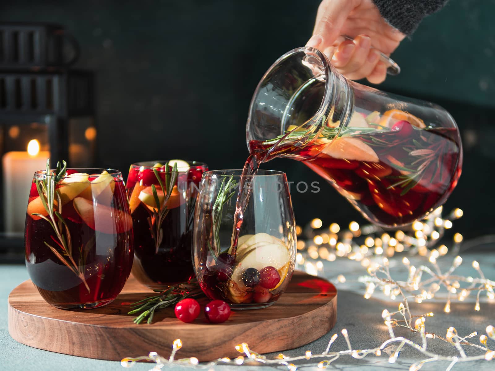 Female hand pours winter sangria in glasses with fruit slice, cranberry and rosemary. Dark christmas holiday background with candle, decoration lighting chain.