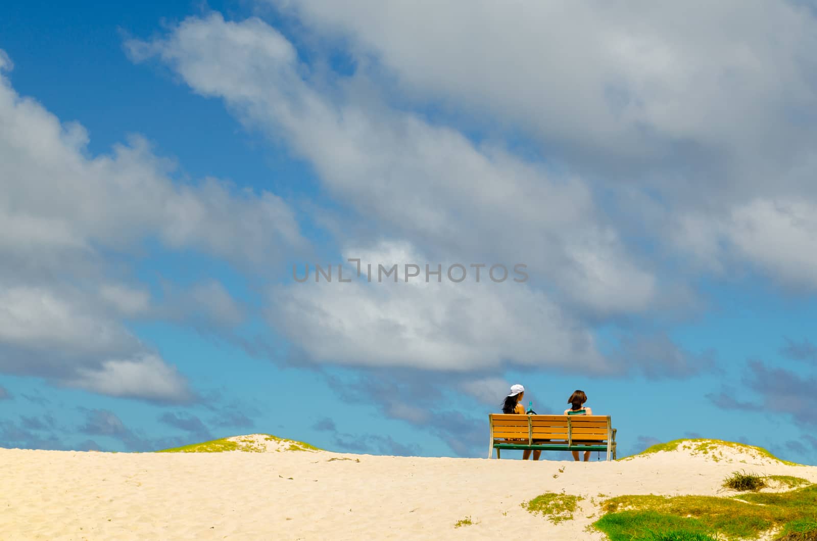 Two girl are sitting on a wooden bank in a Hawaii beach under a cloudy blue sky
