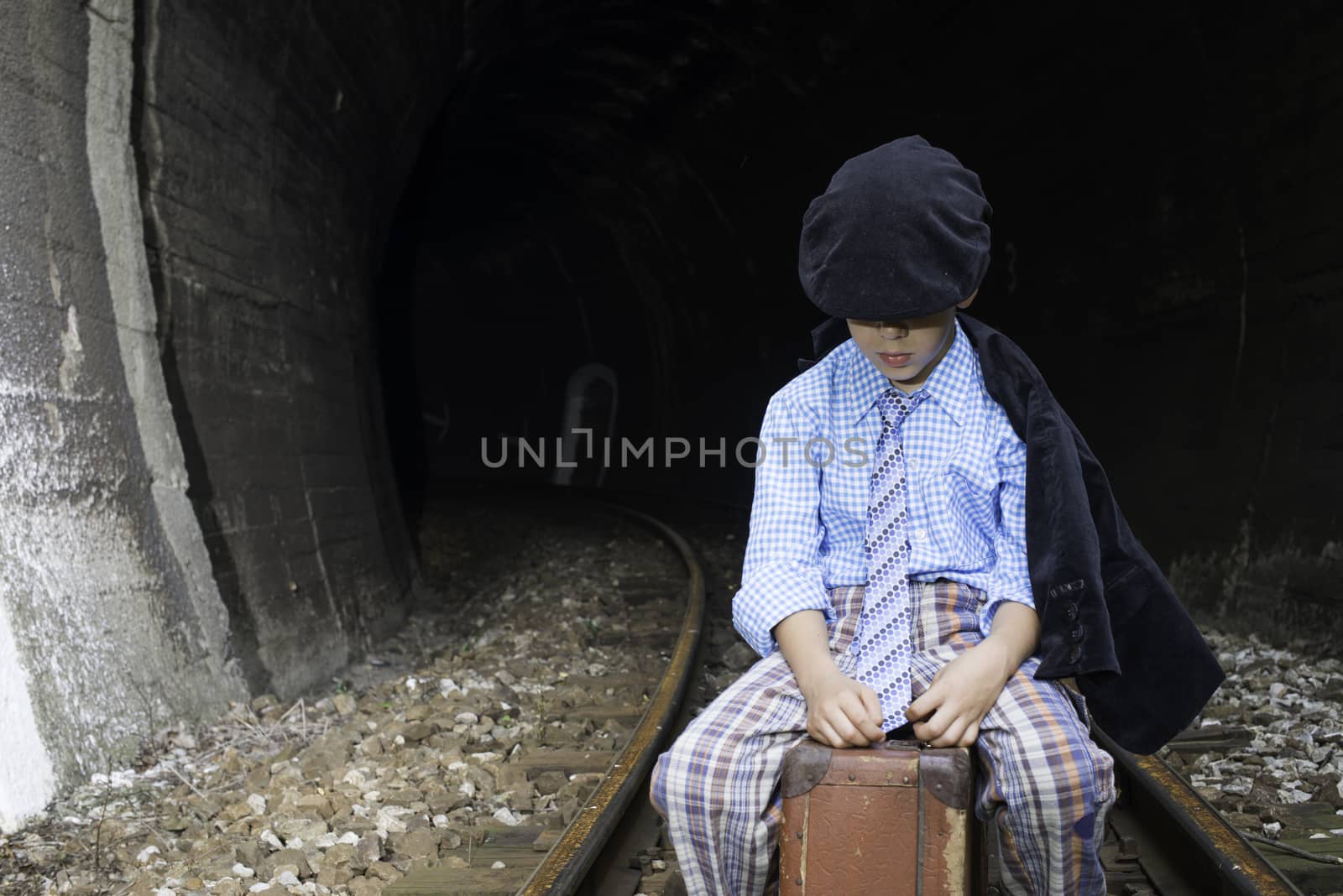 Child in vintage clothes sits on railway road in front of a tunnel.