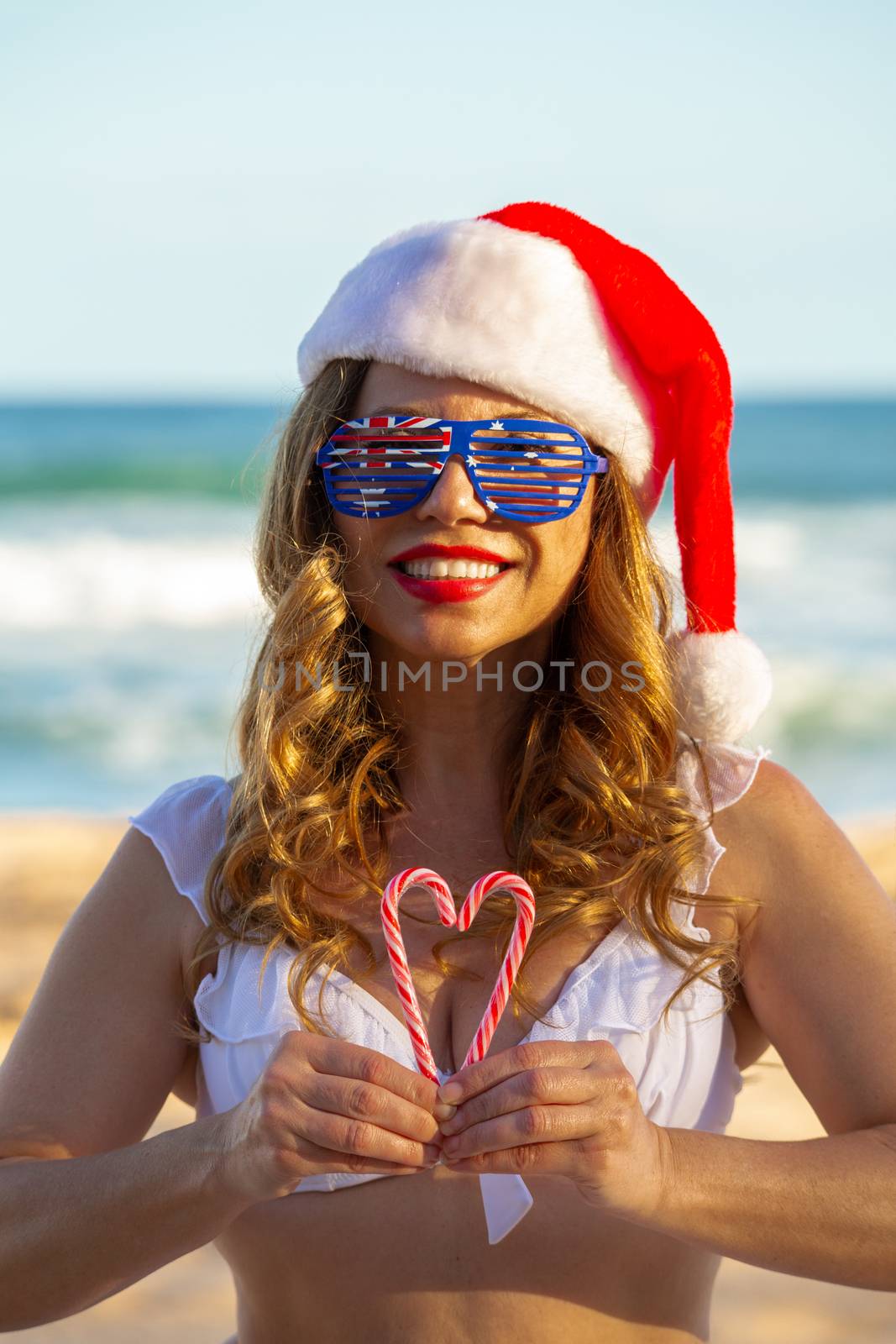 Vibrant smiling woman holds Christmas candy canes in the shape of a heart.   She is at the beach in the glorious summer sunshine
Aussie Christmas