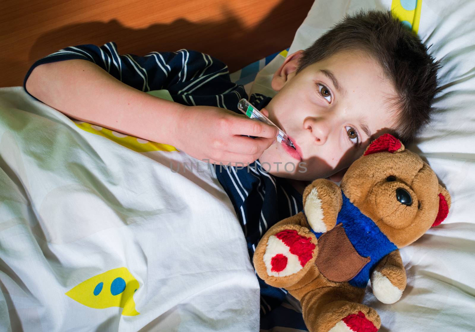 Sick child in bed with teddy bear. Measuring the temperature with a thermometer.