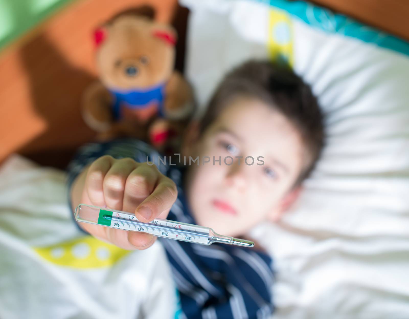 Sick child in bed with teddy bear. Measuring the temperature with a thermometer.