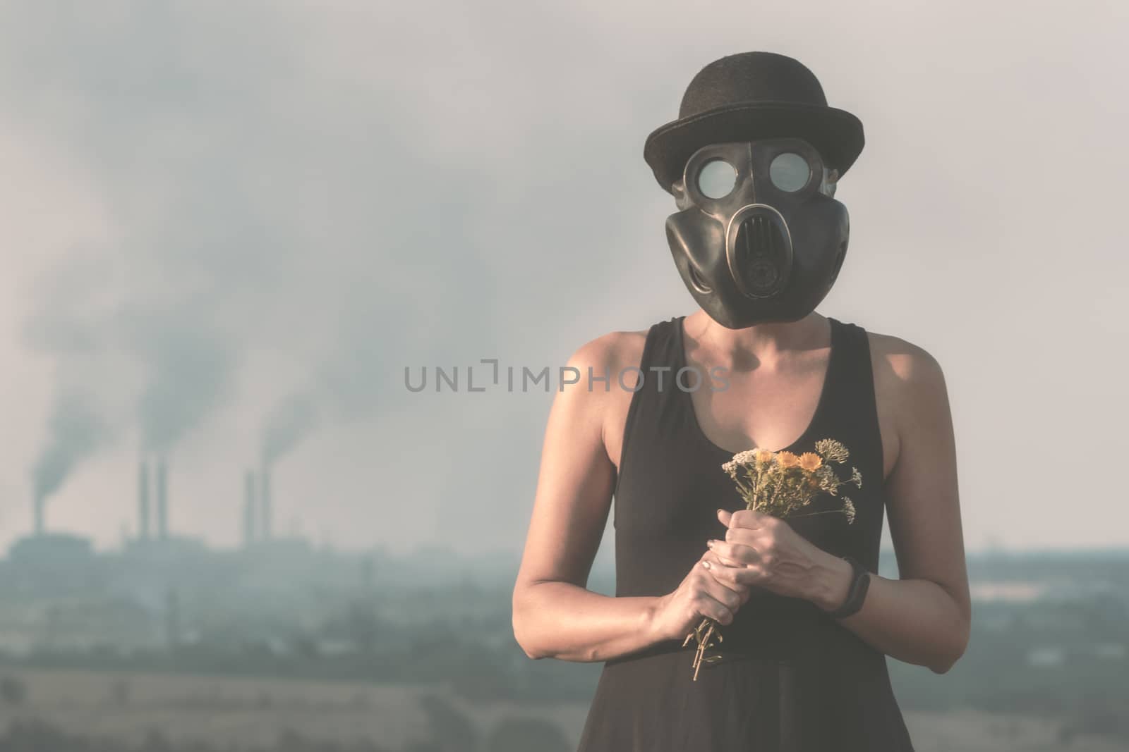 young girl in a black dress and a gas mask with flowers on the background of smoking factory chimneys close up