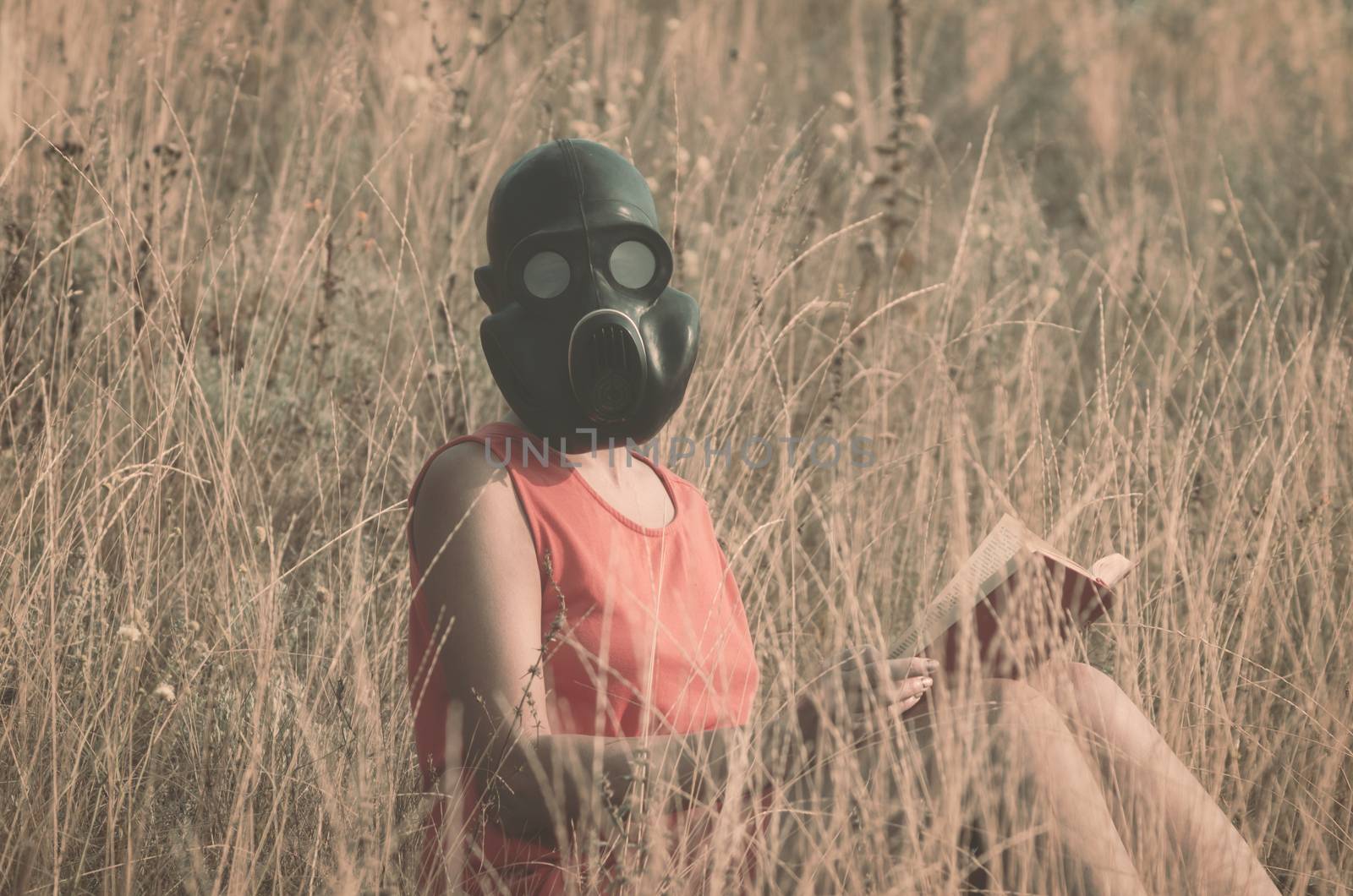 young woman in gas mask in a red dress with a book in her hands sits in the dry grass