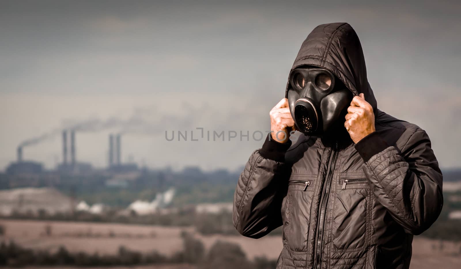 man in a gas mask and a hood against the background of smoke from factory pipes close up