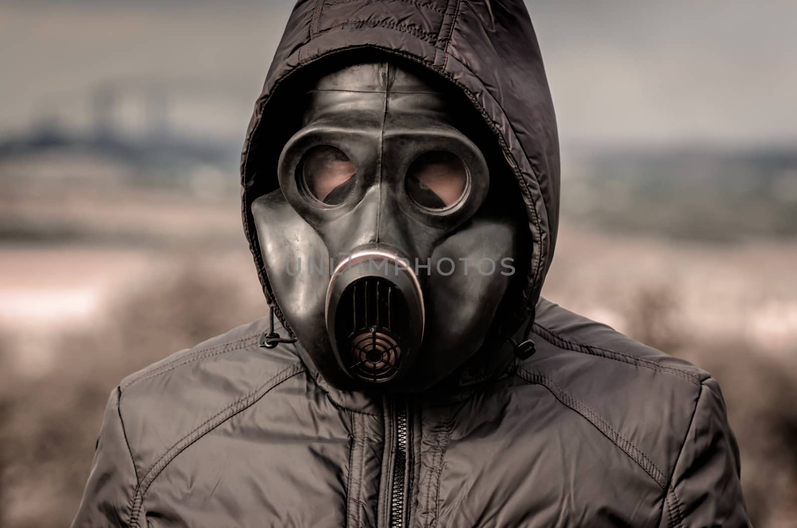 portrait of a man in a gas mask and a hood against the background of smoke from factory pipes close up