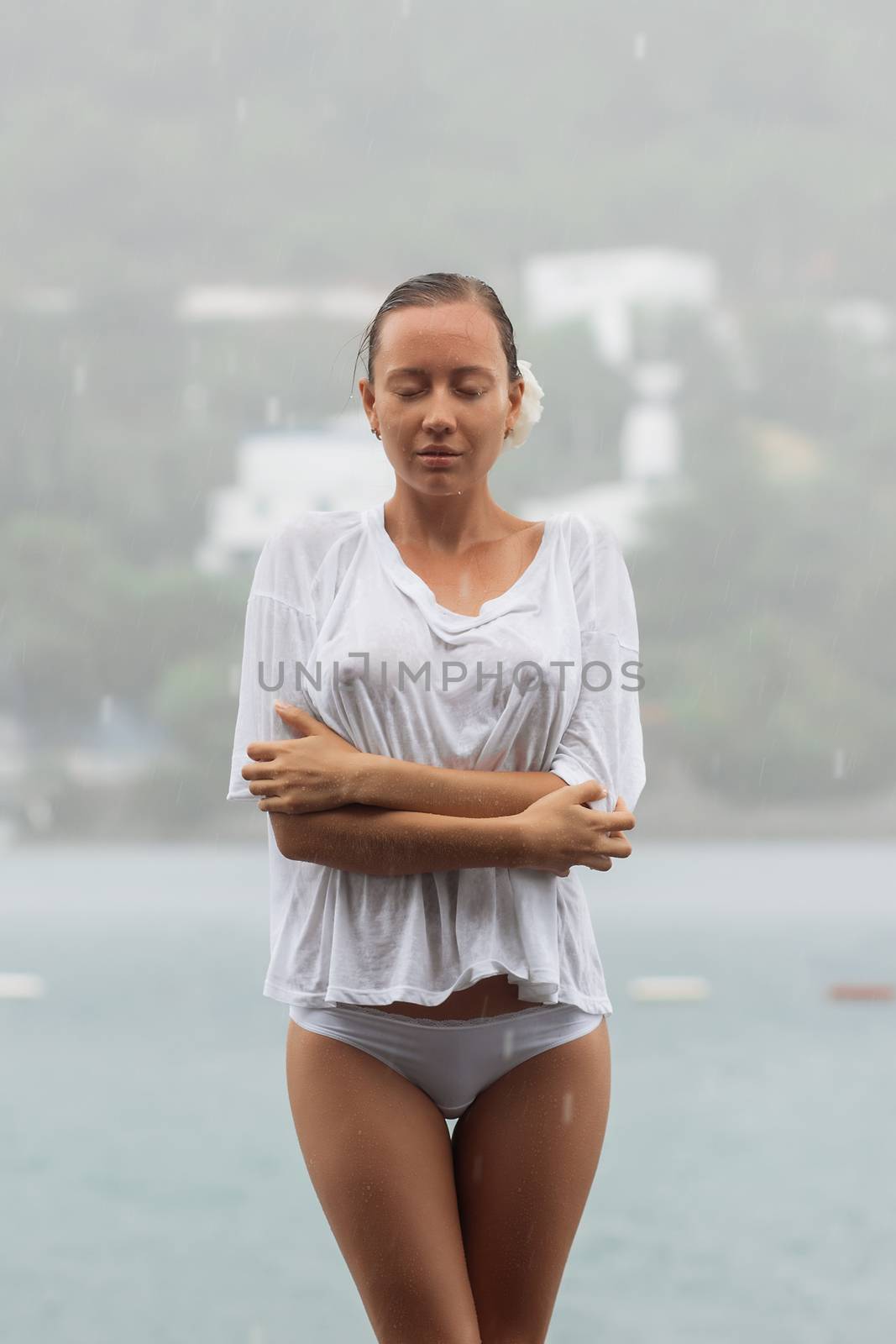 Attractive young female in while blouse and panties embracing herself and keeping eyes closed while standing near water on windy day