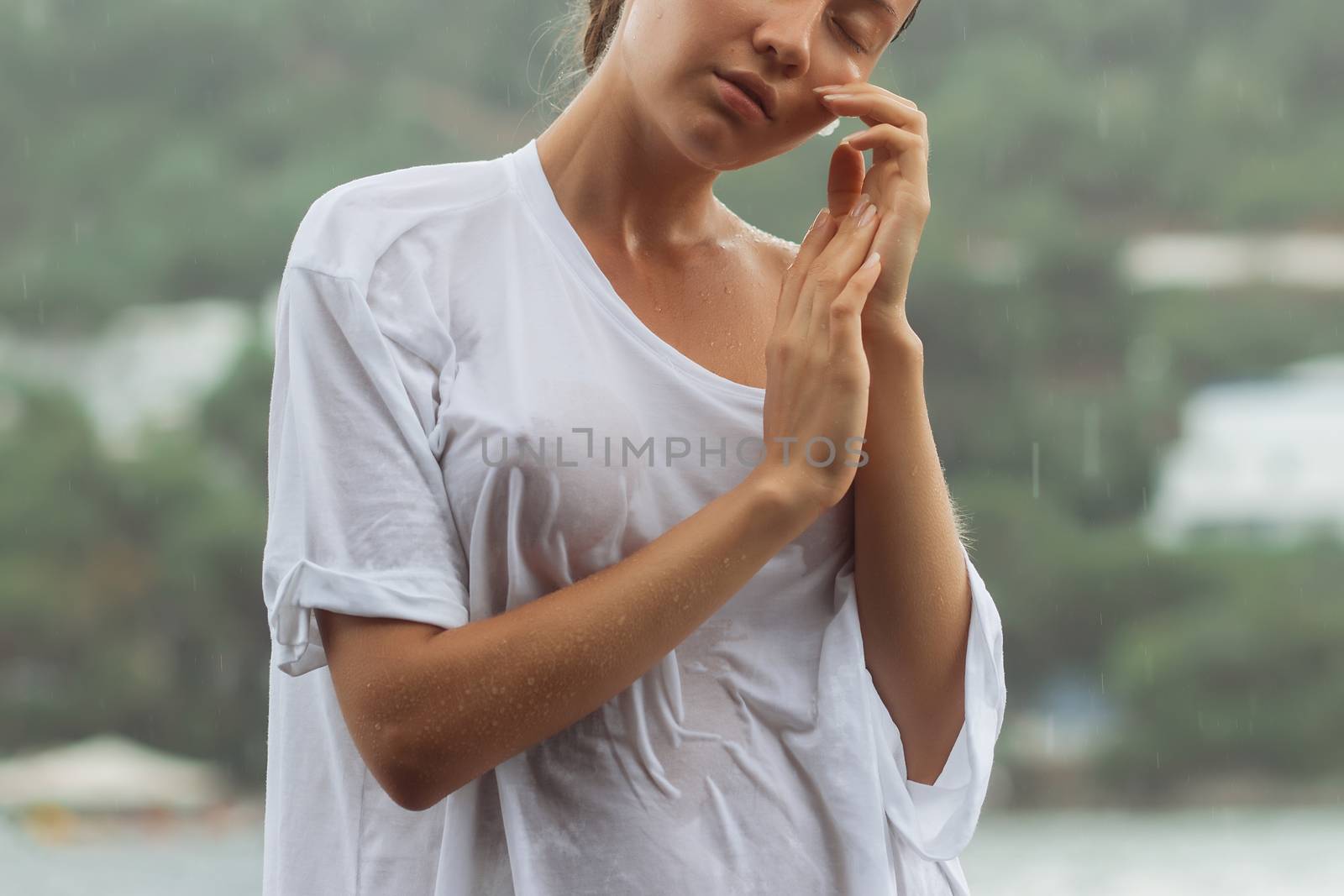 Attractive young female in while blouse and panties embracing herself and keeping eyes closed while standing near water on windy day