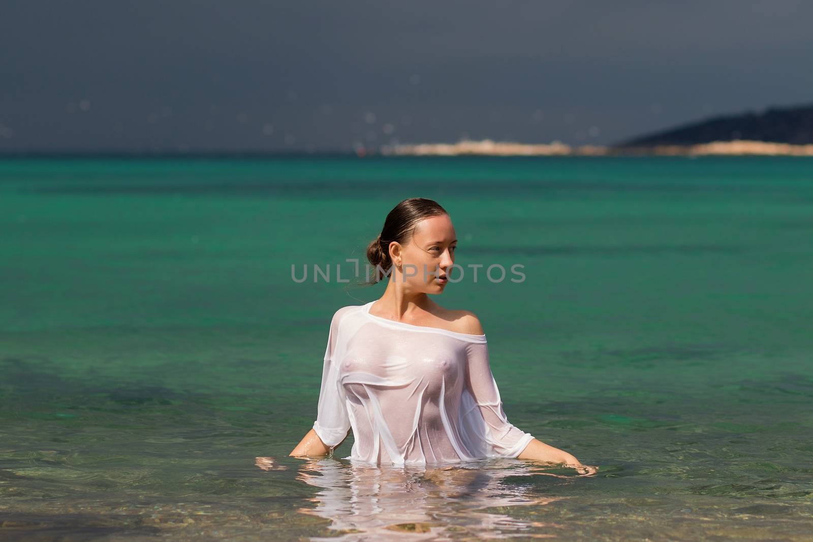 Sensual young female in wet blouse standing in clean sea water and looking away