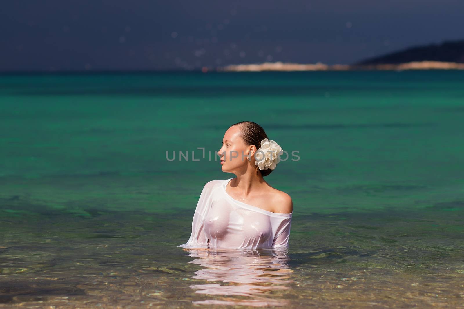 Sensual young female in wet blouse standing in clean sea water and looking away