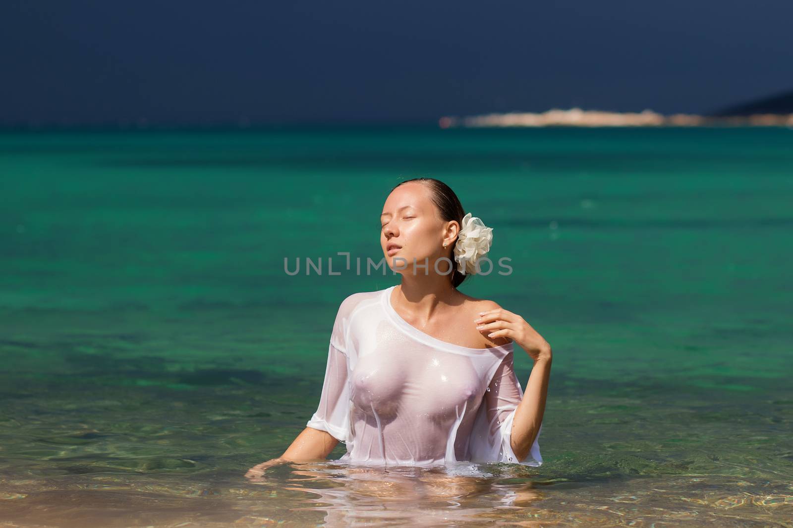 Sensual young female in wet blouse standing in clean sea water and looking away