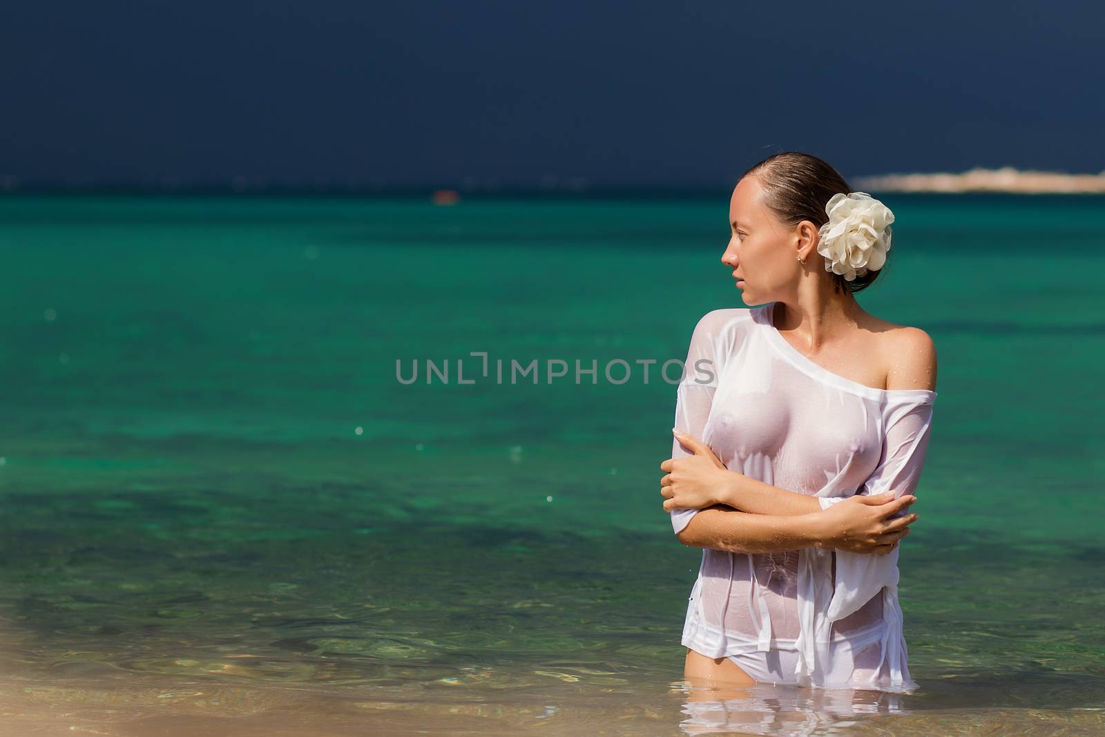 Sensual young female in wet blouse standing in clean sea water and looking away