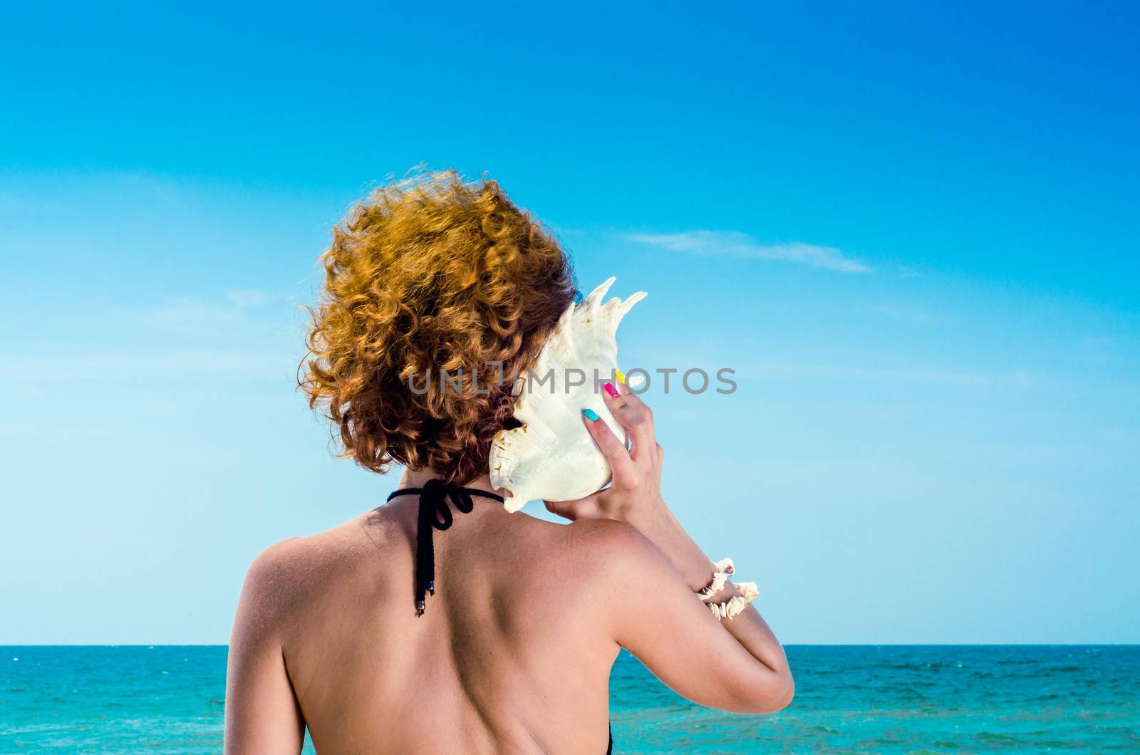 young woman with red hair with a big sea shell and colorful manicure on the background of the sea and blue sky