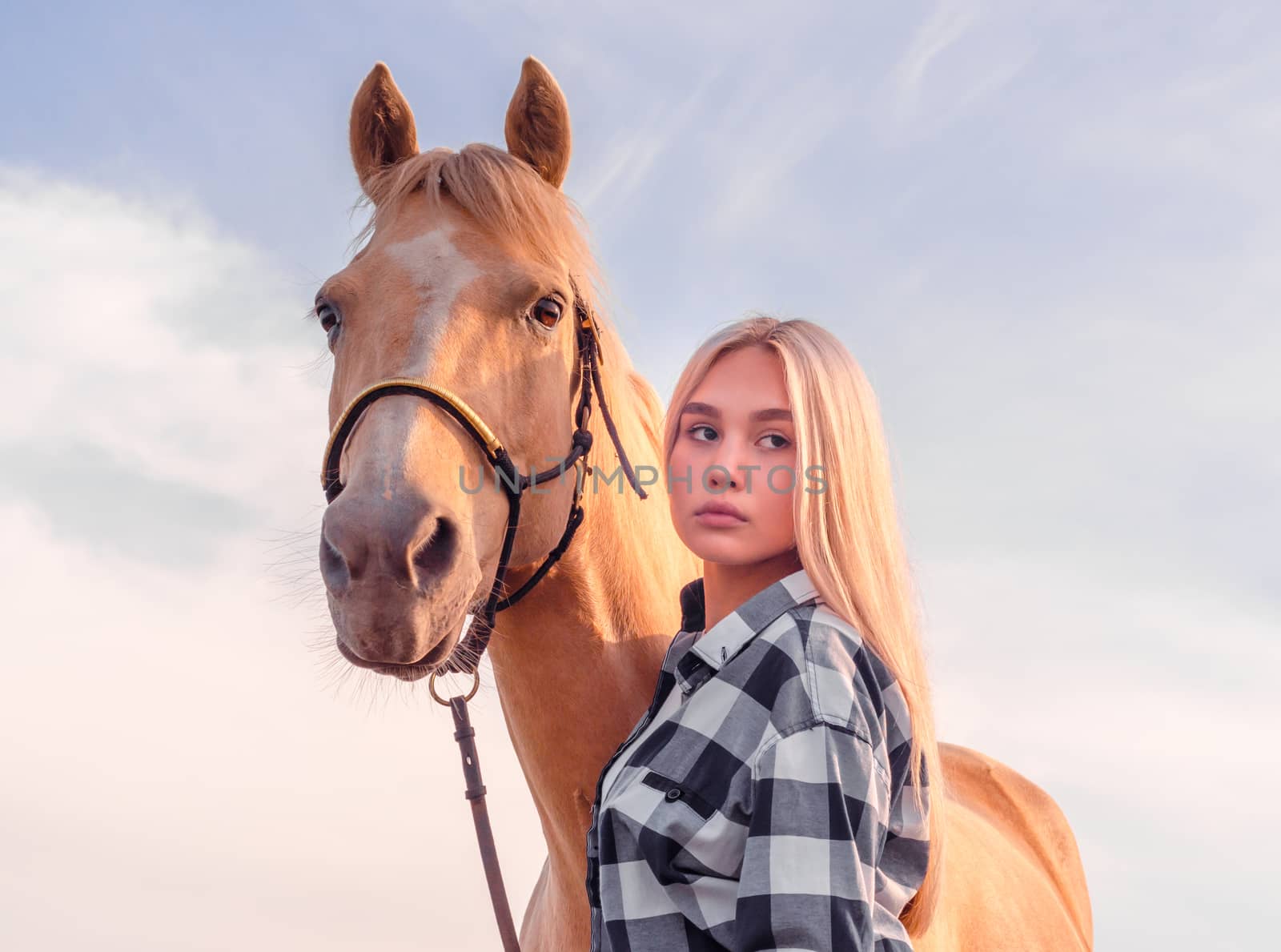 portrait of a young girl blonde with a beige horse against a blue sky with clouds