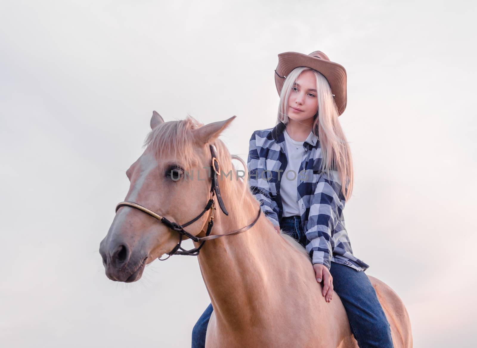 young girl dressed in a cowboy hat and blue jeans sits on a beige horse