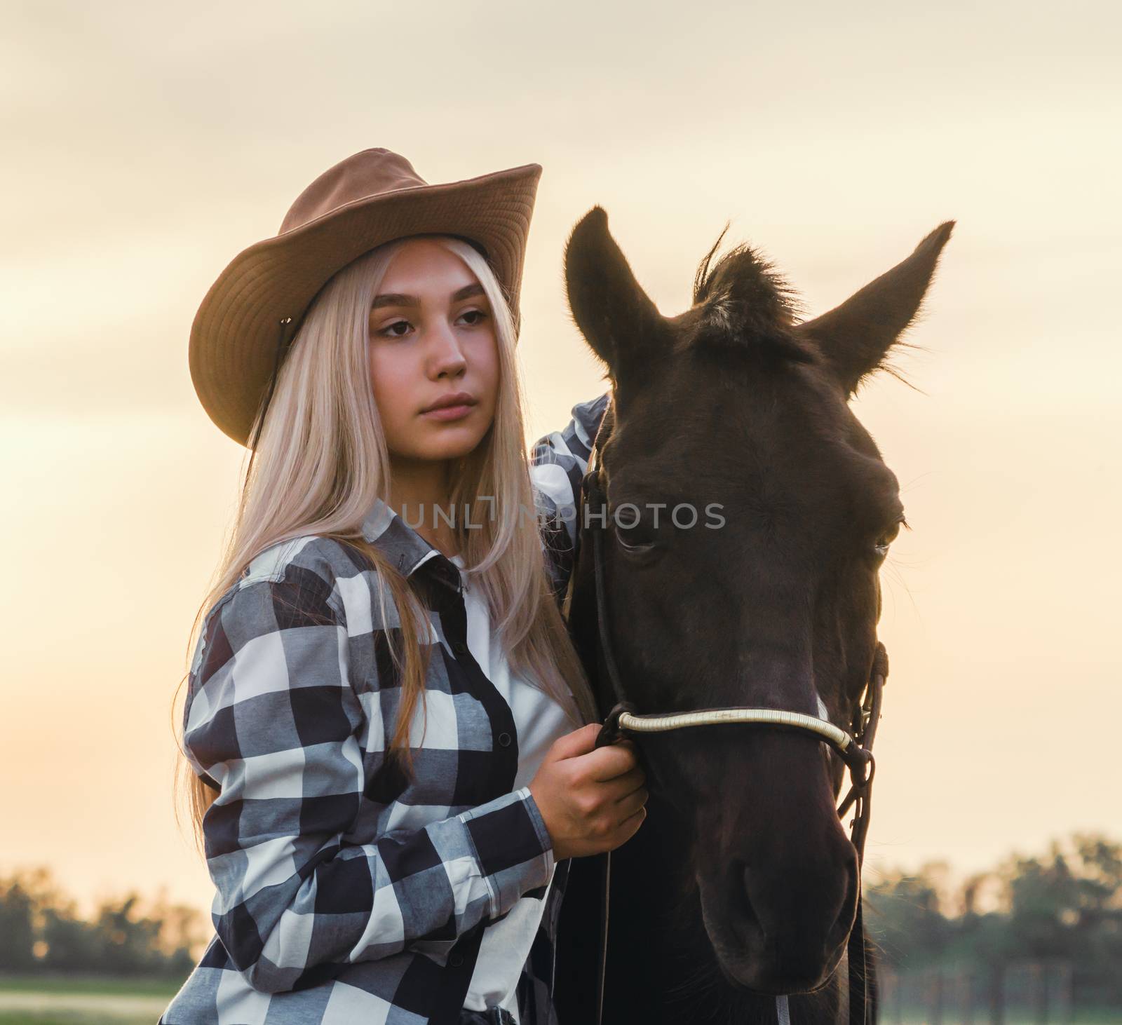young girl dressed in a cowboy hat and a plaid shirt with a horse at sunset
