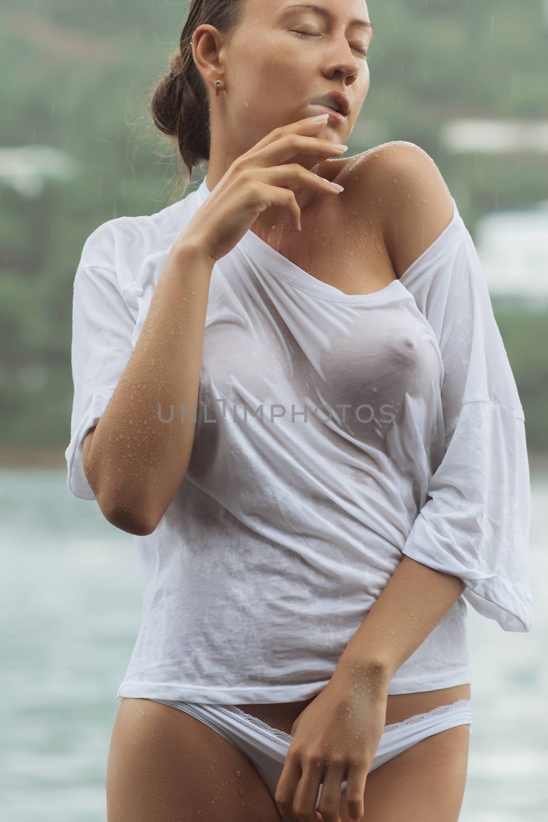 Attractive young female in while blouse and panties embracing herself and keeping eyes closed while standing near water on windy day
