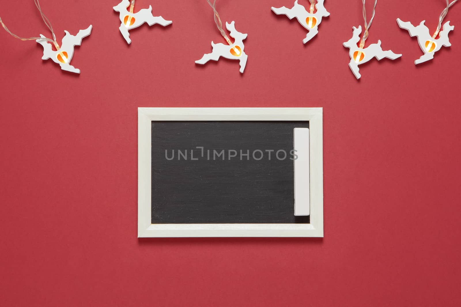 Christmas garland in form of white running deers, small chalk board with copy space on dark red background. Blank for postcard layout. Festive, New Year concept. Horizontal, flat lay. Minimal style.