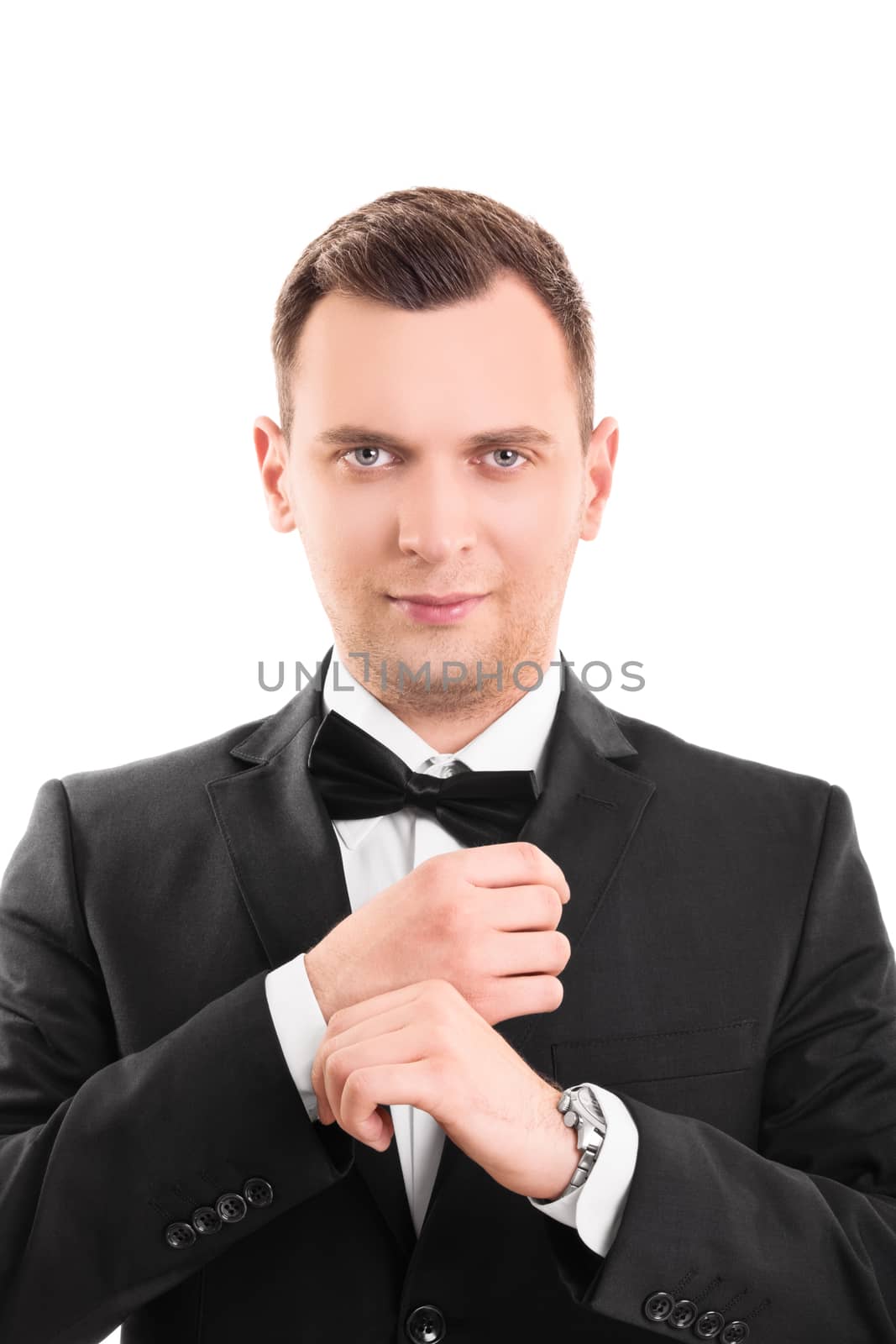 Close up portrait of a confident, stunning, trendy, attractive, dreamy, perfect man in black suit with bow tie, adjusting the cufflinks on his white shirt, looking at the camera, isolated on a white background.