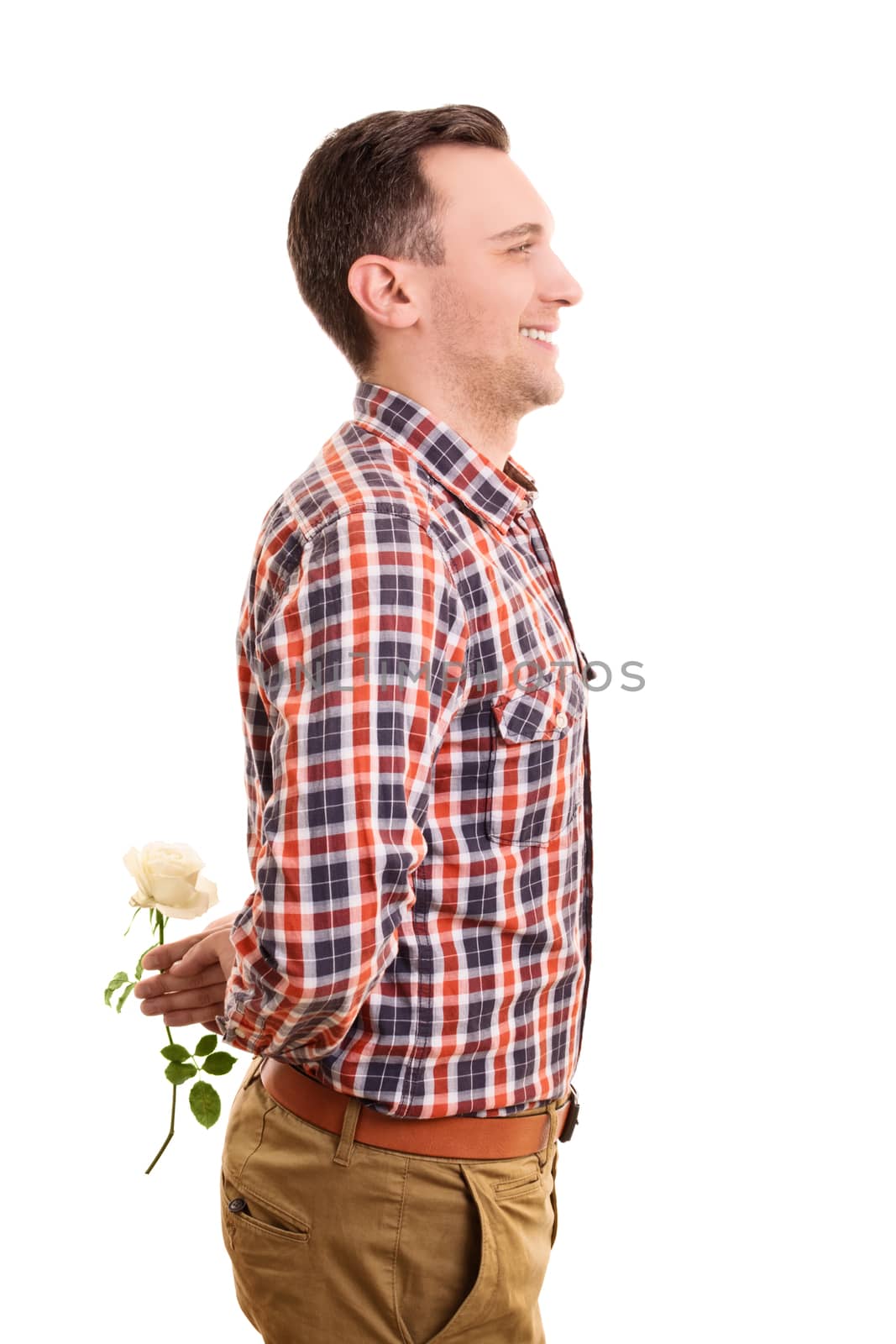 Valentine's Day concept. Portrait of a romantic stylish handsome young man hiding a white rose flower behind his back, standing and smiling, isolated on white background.