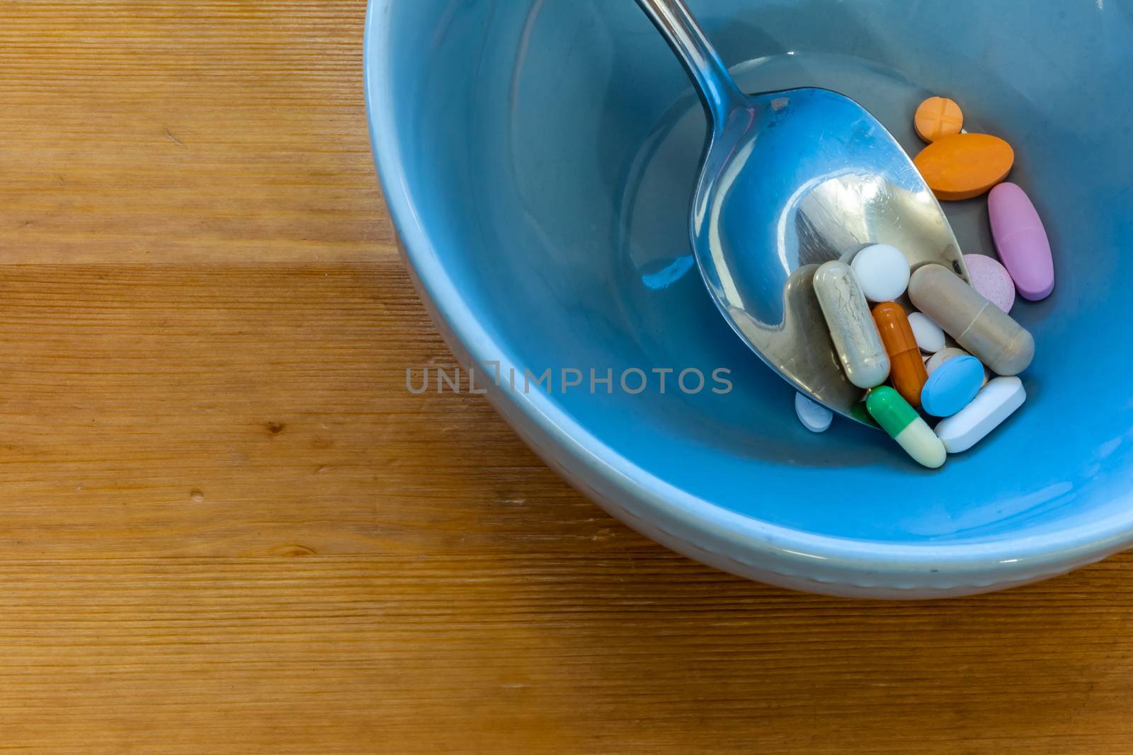 A collection of pills, including pharmaceutical drugs, vitamin tablets, over-the-counter medications and herbal supplements, appears in a soup bowl on a wooden table with a metal spoon.