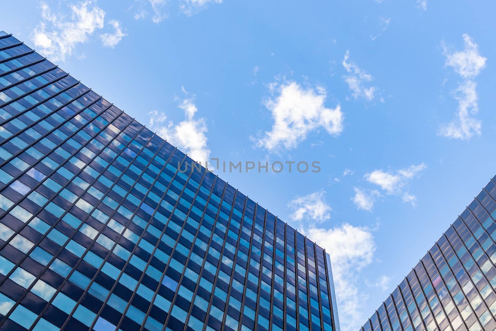 Commercial office buildings are viewed from the ground, looking up. A blue sky with a scattering of white clouds can be seen above, and in the reflecting windows. Their roofs of angle in and down.