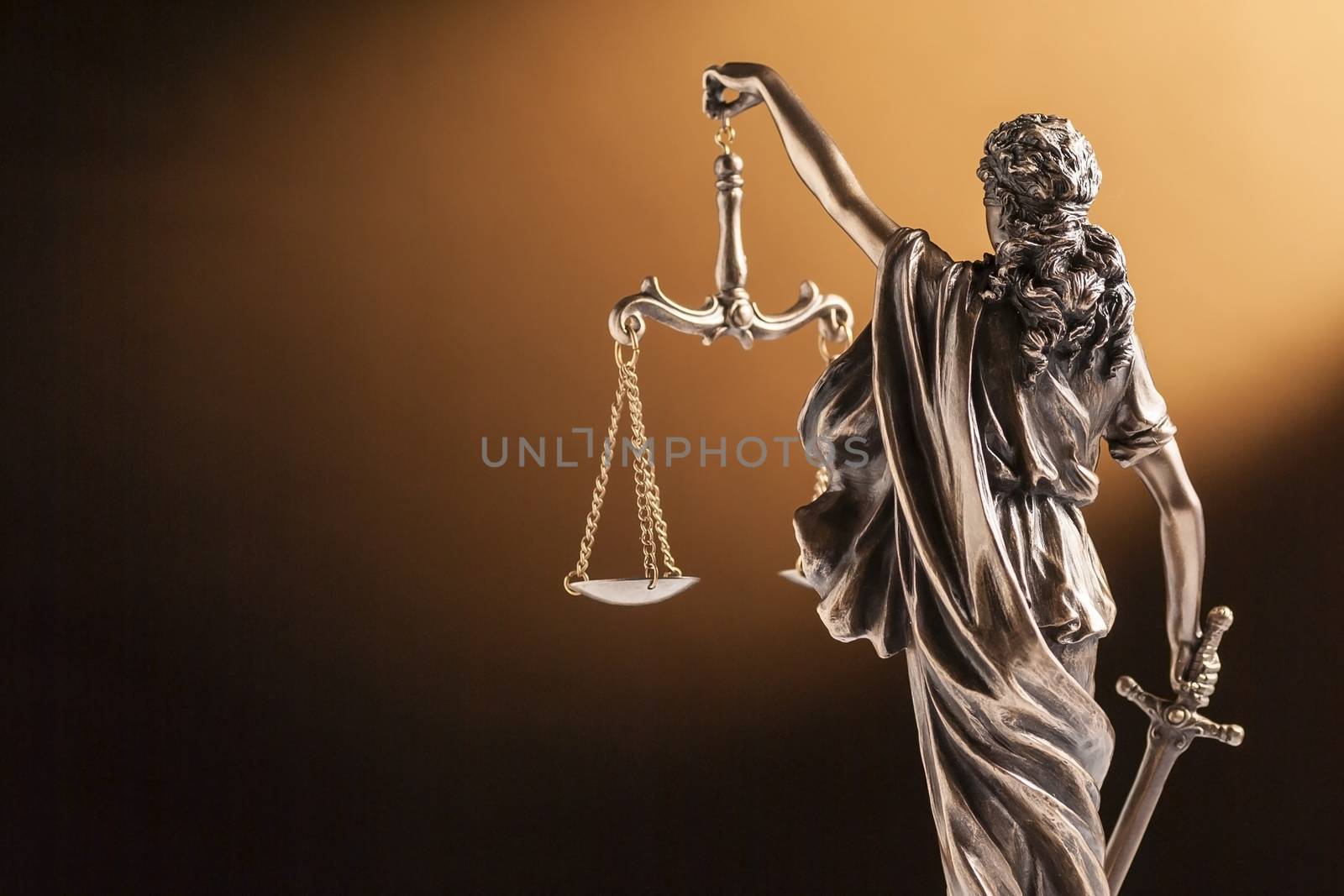Rear view of of a small bronze figurine of Justice holding up the scales of law and order and carrying a sword facing towards blank copy space on brown