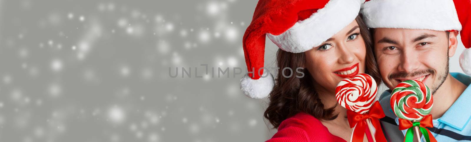 Young happy smiling couple in christmas santa hats with lollipops on magic snow bokeh background with copy space