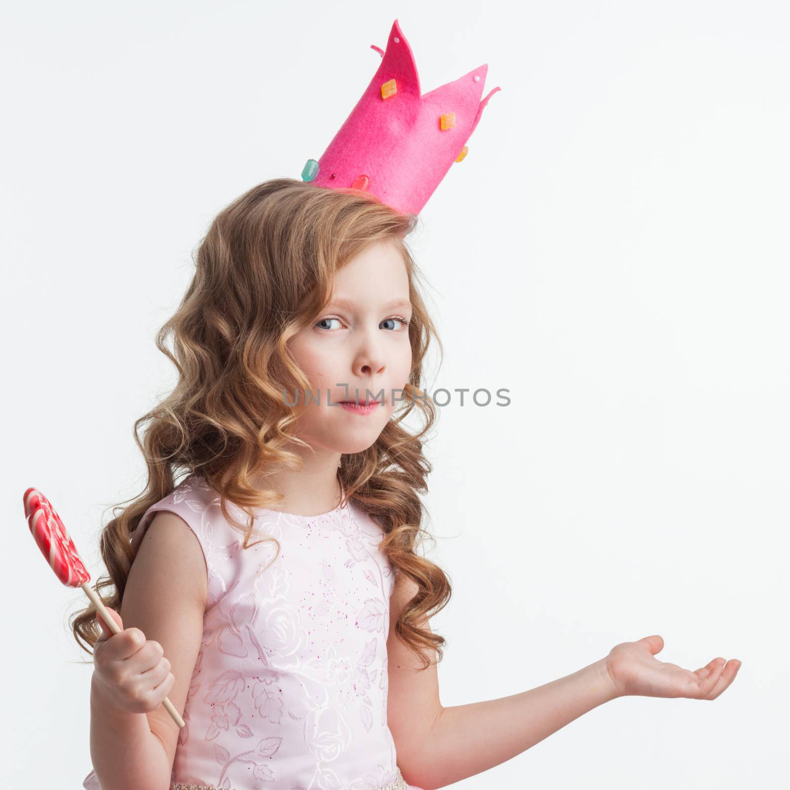 Beautiful little candy princess girl in crown holding big lollipop and posing with hands up in air saying, why, i dont know, so what isolated on white background