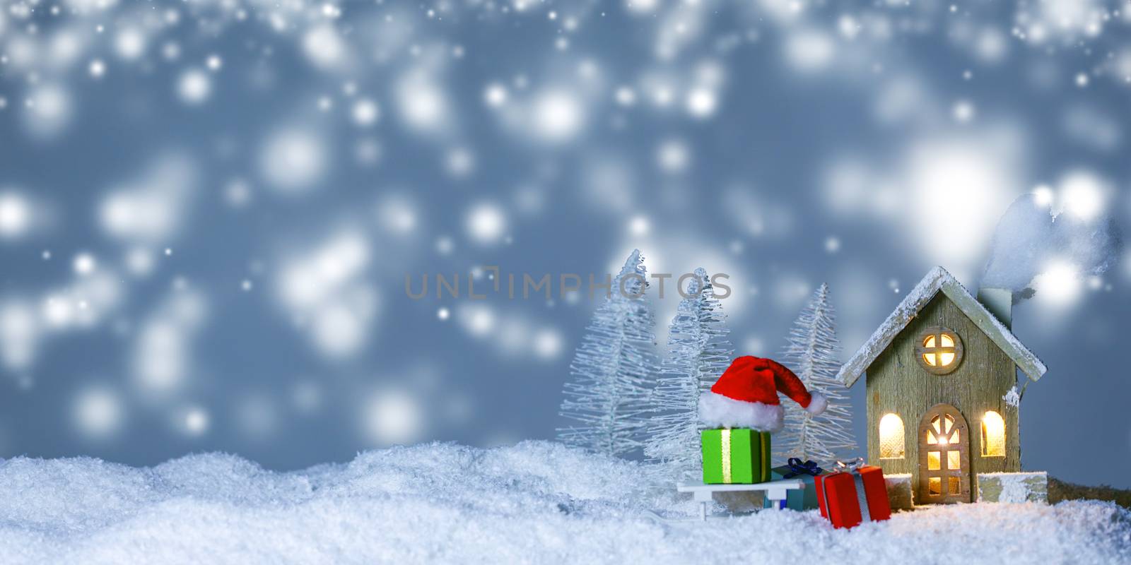 Christmas card with small glowing decorative house , gifts and Santa Claus cap on sleigh on snow over fairy tale winter fir forest background and falling snow . New Year design postcard . Copy space for text